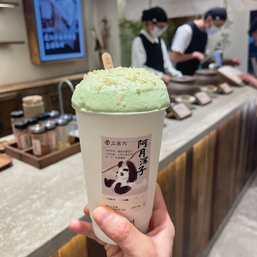 A hand holds a paper cup of tea topped with pistachio foam, while in the background members of staff work at a counter, at Li Shanshan Tea café.